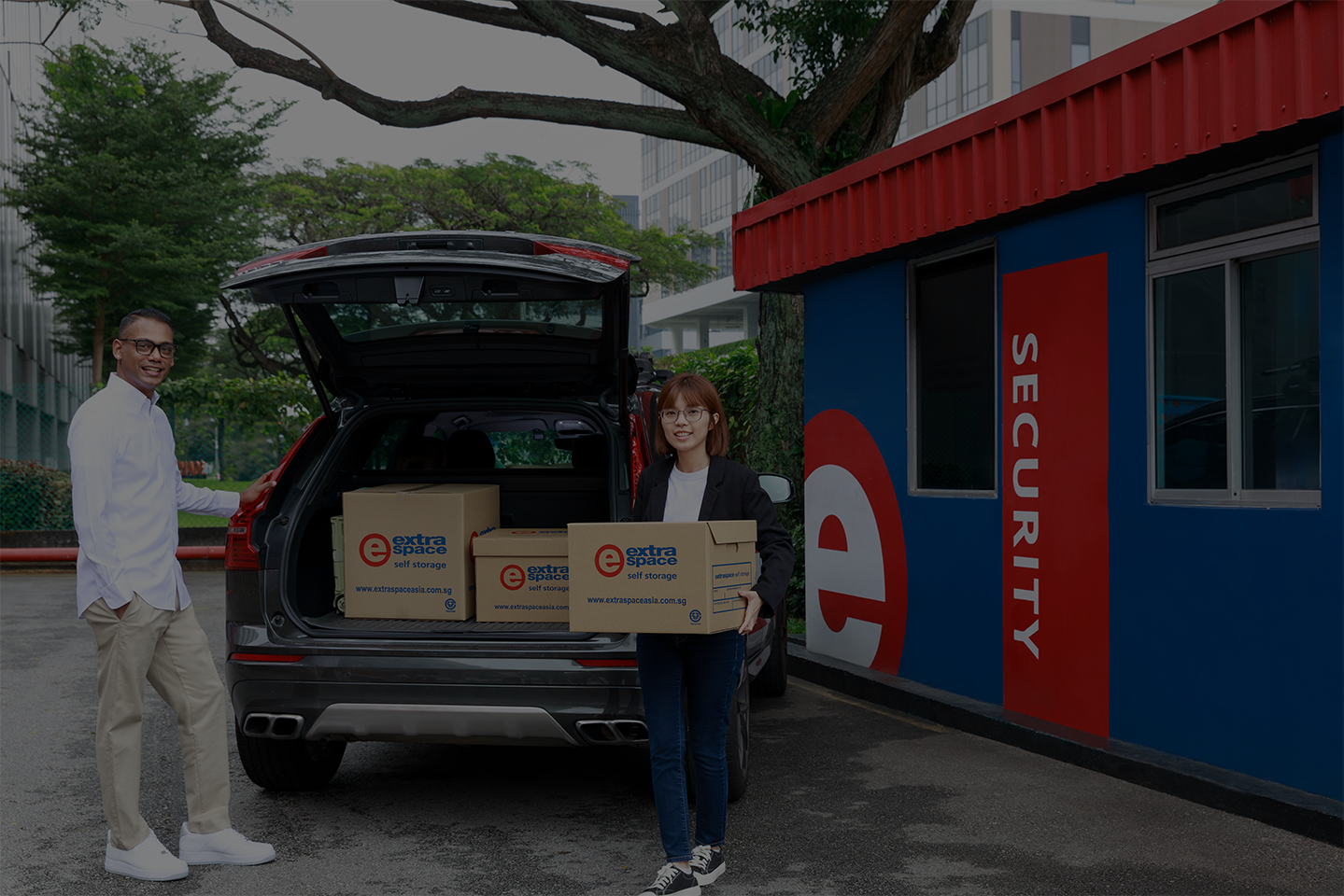 Two people unloading Extra Space storage boxes from a car outside a security building.